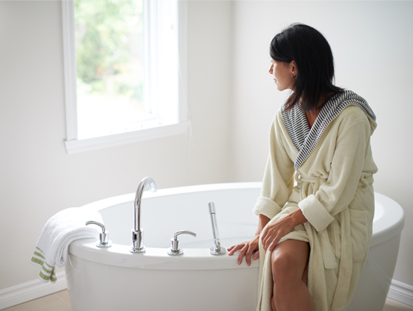 A woman wearing a bathrobe sits on the side of a white elliptical-shaped soaker tub and looks out a window