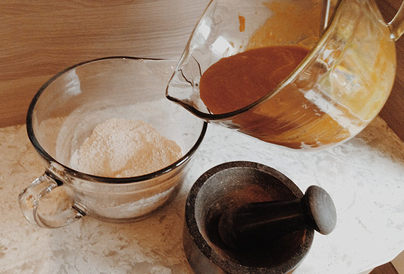 A glass measuring cup filled with pumpkin mixture is about to be poured into a glass measuring cup filled with flour