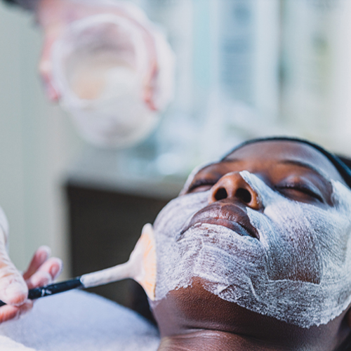 A woman lying down on a spa bed while a person brushes a chemical peel treatment on her face.