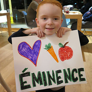 a young child holds a hand drawn sign depicting a heart, a carrot, an apple and the word Eminence