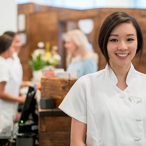 Smiling esthetician stands near the front desk where staff are assisting a client.